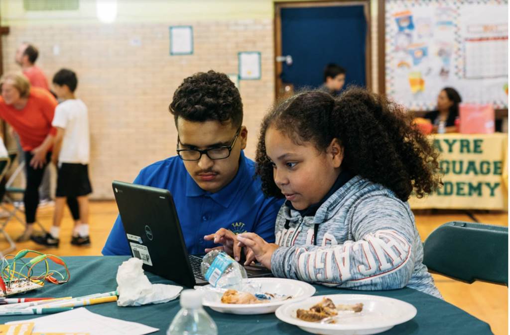 A man sits with a student using a laptop